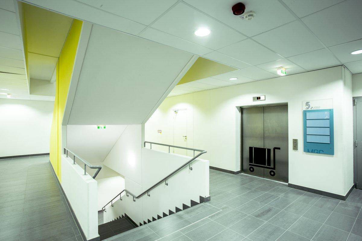 Helles Treppenhaus mit gelben Akzenten, Fliesenboden und einem modernen Aufzug im Hintergrund.