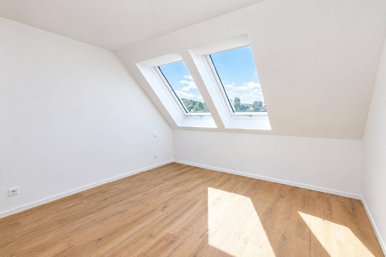 Helles Zimmer im Dachgeschoss mit zwei Dachfenstern, Holzboden und weißen Wänden.