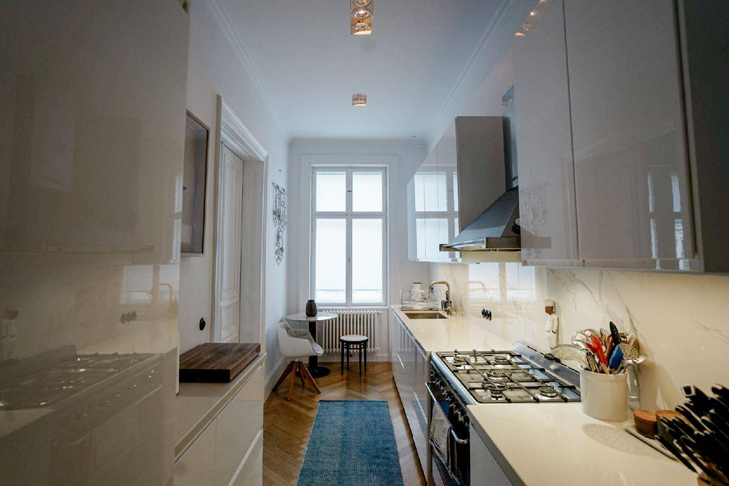 Schmale, moderne Küche mit weißen Hochglanzschränken, Gasherd und Fenster.