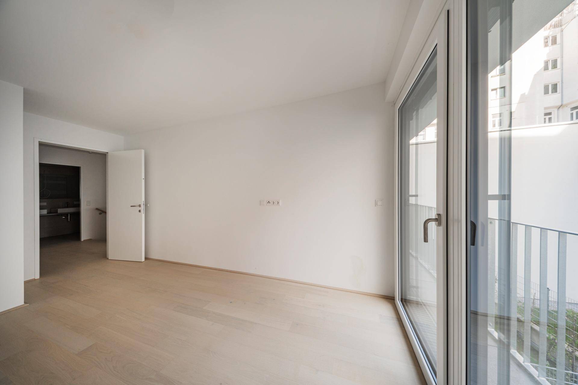 Helles Zimmer mit Holzboden und großem Fenster, das zu einem französischen Balkon führt.
