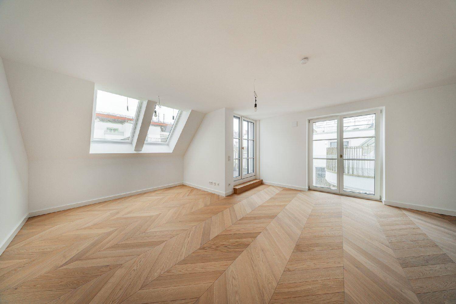 Lichtdurchfluteter Raum mit Fischgrätparkett, zwei Dachfenstern und Zugang zu einem Balkon.
