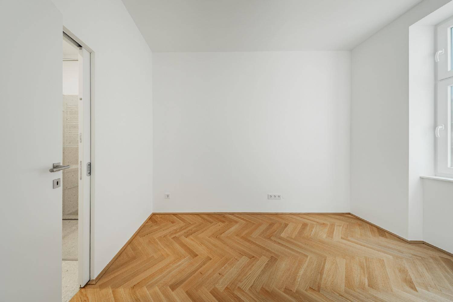 Leeres Zimmer mit Fischgrätparkett und Zugang zu einem angrenzenden Badezimmer, hell und frisch renoviert.