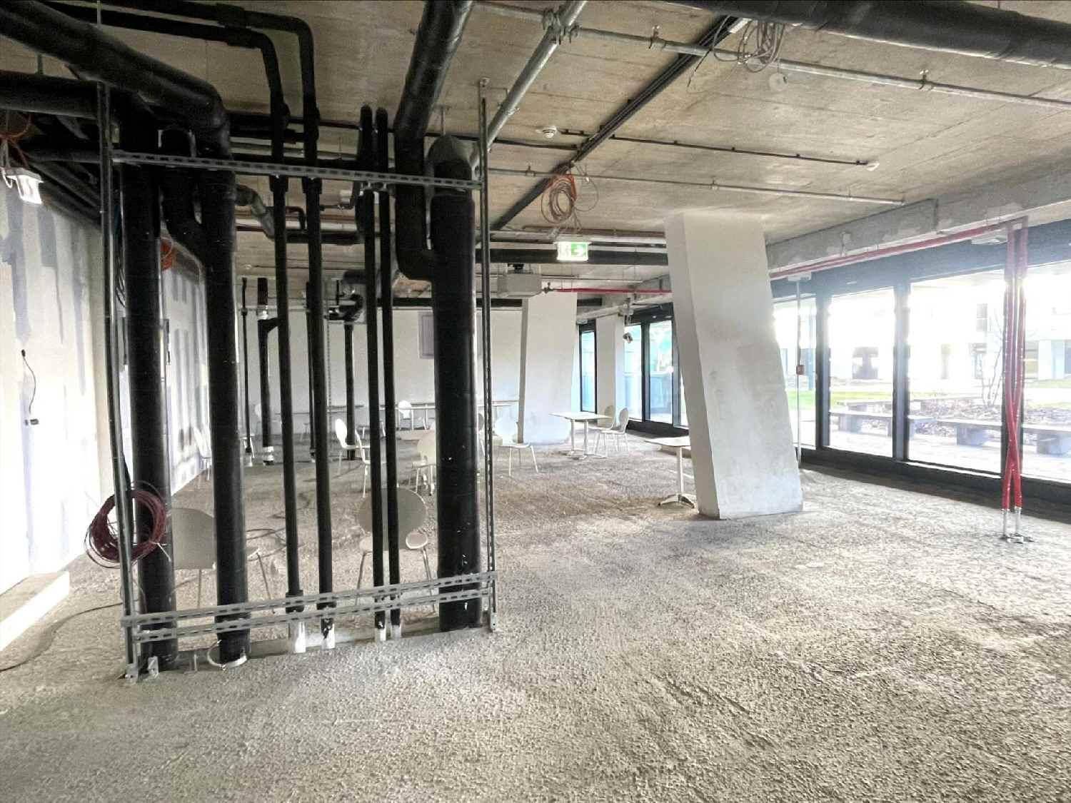 Unfertiger Gewerberaum mit freiliegenden Rohren und Betonstruktur, viel Tageslicht durch große Fenster.
