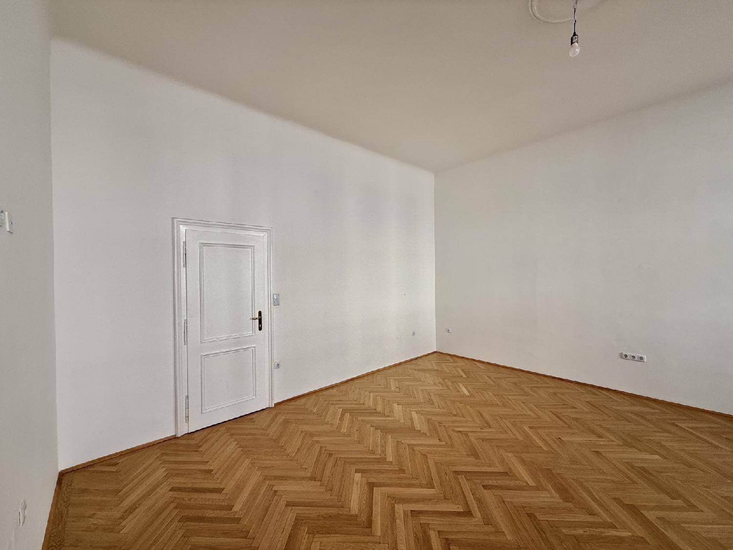 Eckbereich eines leeren Zimmers mit Fischgrätparkettboden und einer klassischen weißen Tür, bereit zur Einrichtung.