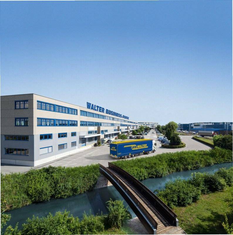 Modernes Bürogebäude eines Geschäftsparks mit LKW-Zufahrt und einem kleinen Fluss im Vordergrund.