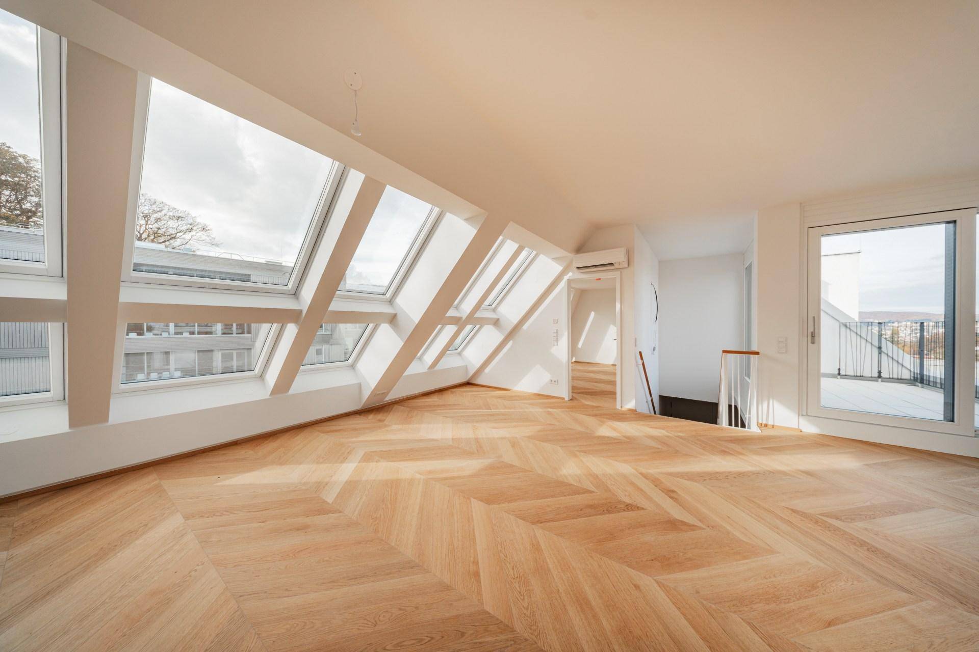 Helles Dachgeschosszimmer mit modernem Fischgrätparkett, großen Dachfenstern und Zugang zu einer Terrasse.