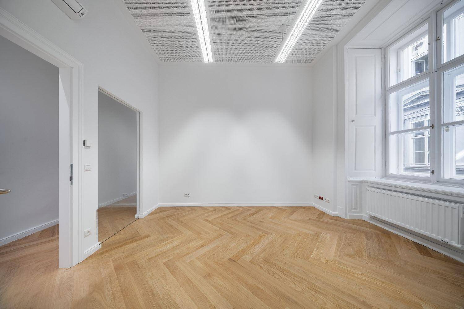 Heller, leerer Raum mit Fischgrätparkett, großem Fenster mit Fensterläden und Klimaanlage.