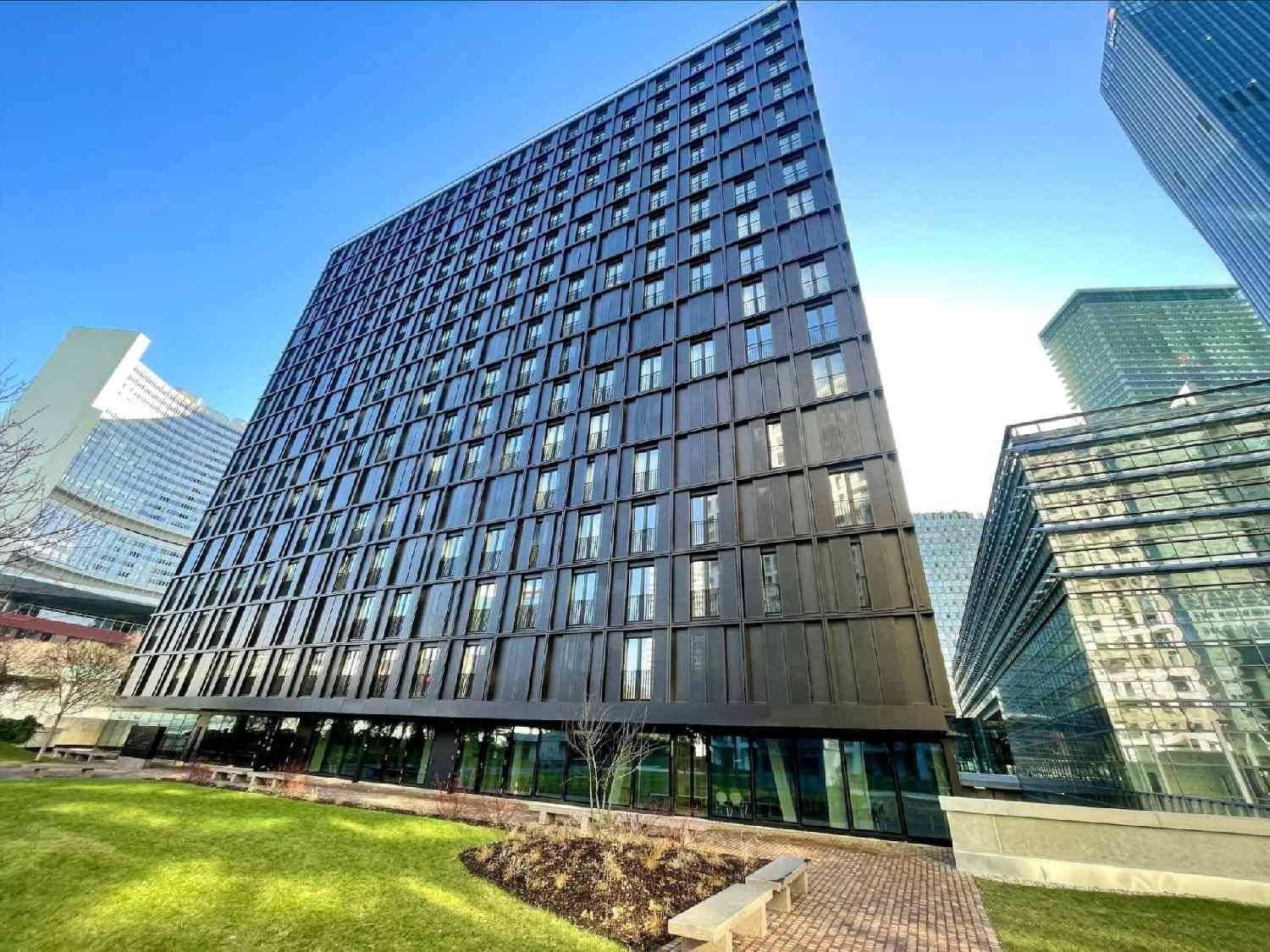 Hohes, modernes Gebäude mit dunkler Fassade und vielen Fenstern, umgeben von einer gepflegten Grünfläche unter blauem Himmel.