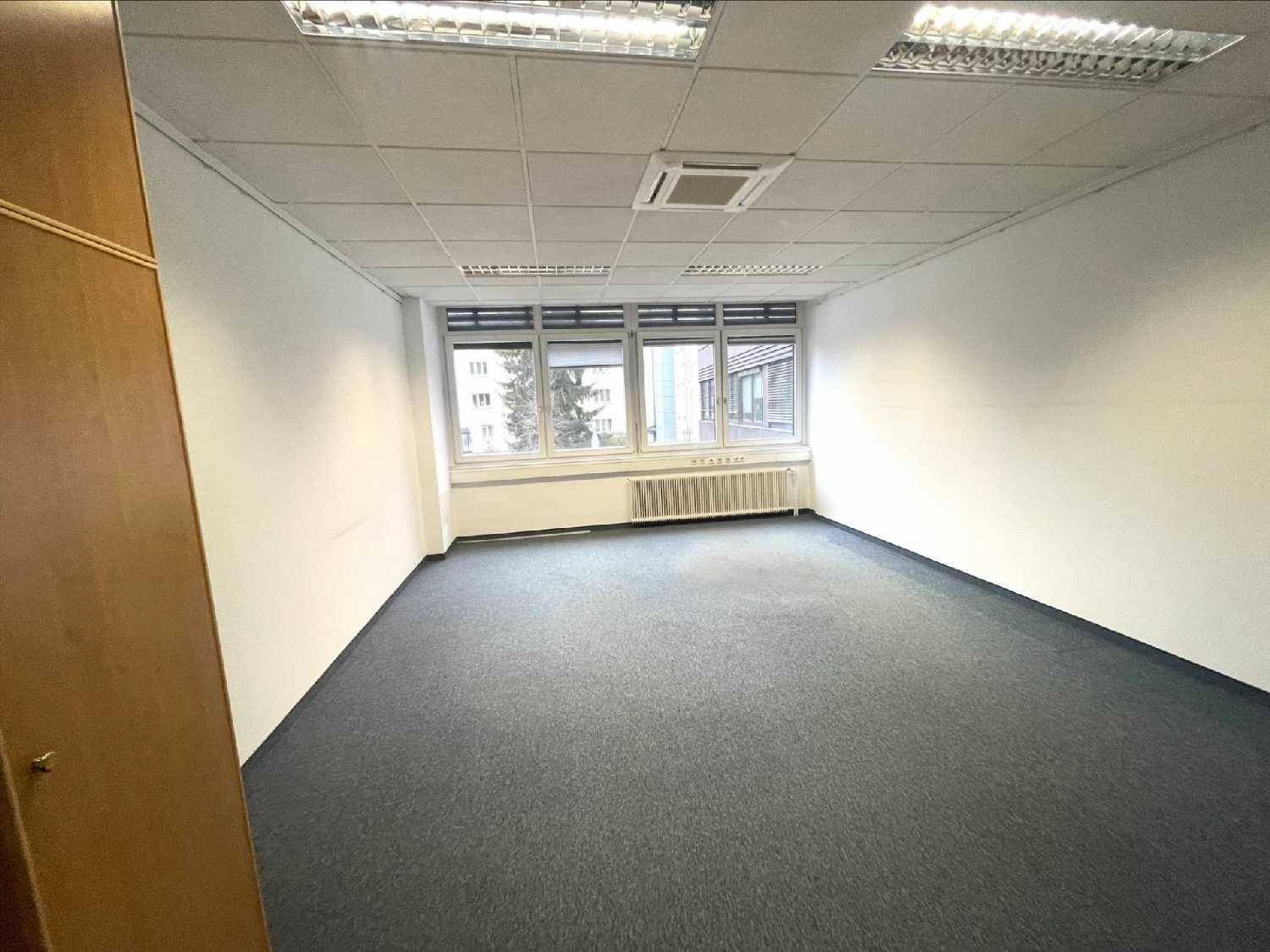Leeres Büro mit grauem Teppichboden, weißen Wänden und Fenstern, teilweise sichtbar ein Holzschrank.