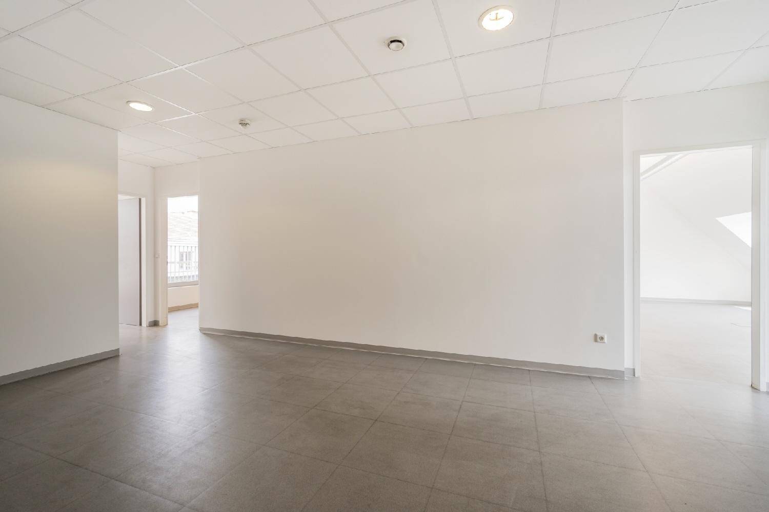 Offener Bürobereich mit weißen Wänden, grauen Fliesen und Zugang zu einem weiteren Raum.