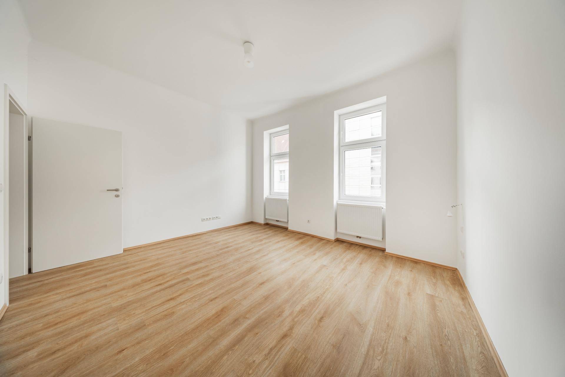 Lichtdurchfluteter Raum mit hellem Laminatboden und zwei Fenstern, ideal für ein Wohnzimmer.