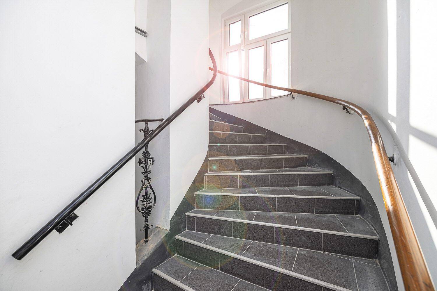 Geschwungene Treppe mit Fliesenstufen und Holzgeländer in einem historischen Gebäude.