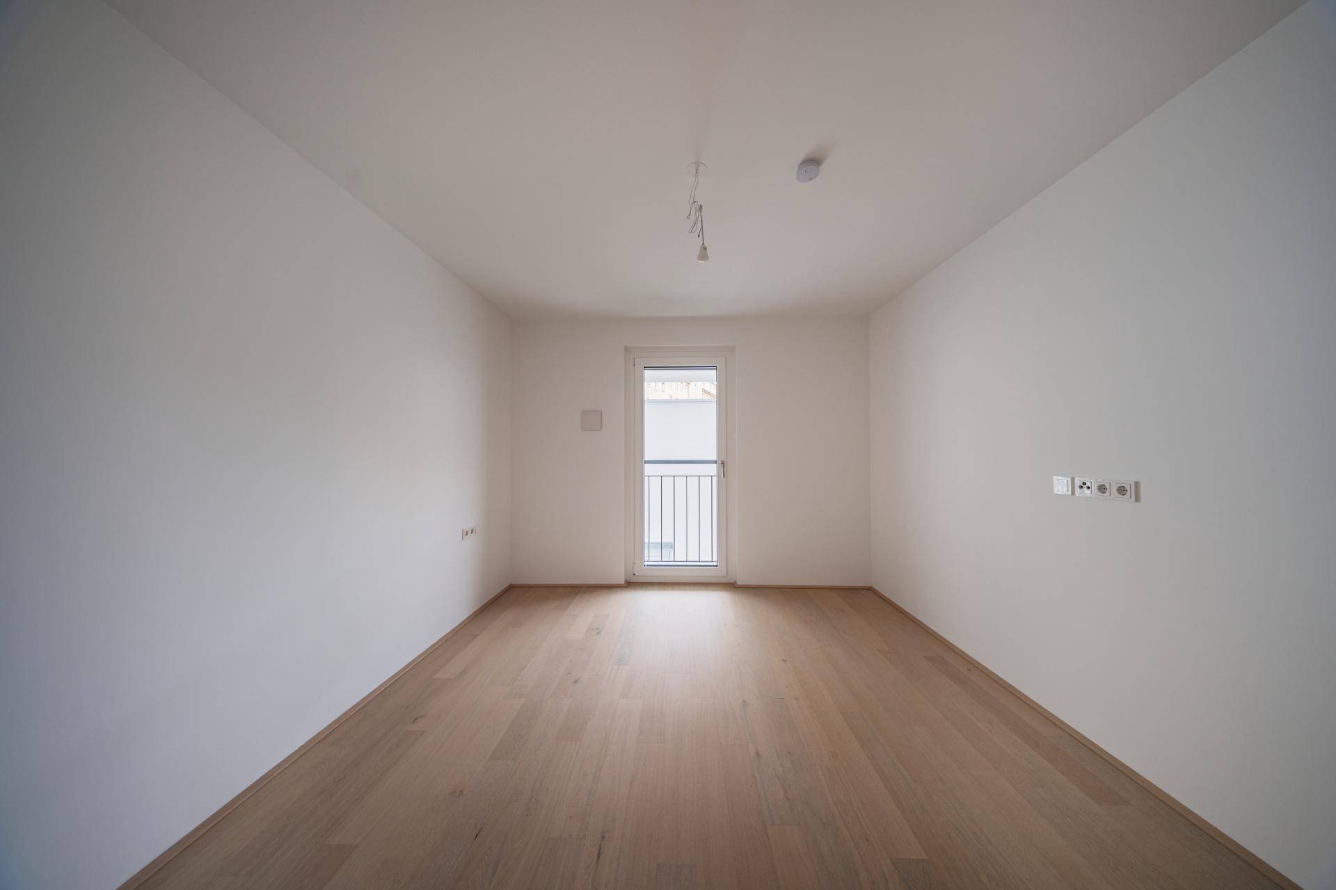 Leeres Zimmer mit hellem Parkettboden und einem französischen Balkon, der viel Licht hereinlässt.