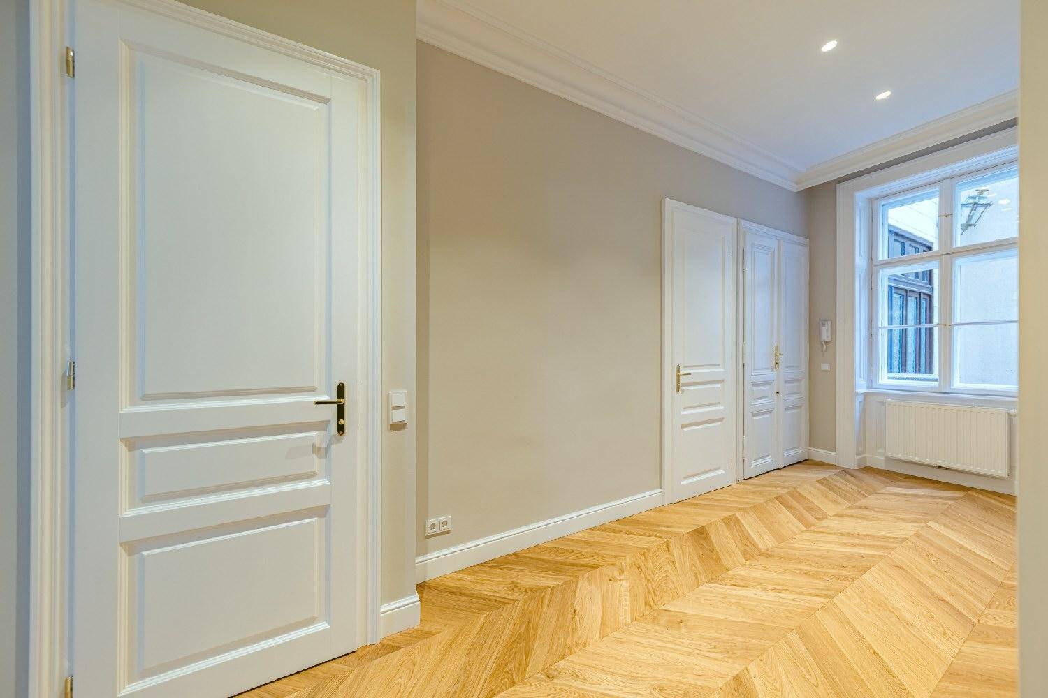 Flur mit Fischgrätparkett, weißen Türen und Einbauschränken, der zu einem hellen Zimmer mit Fenster führt.