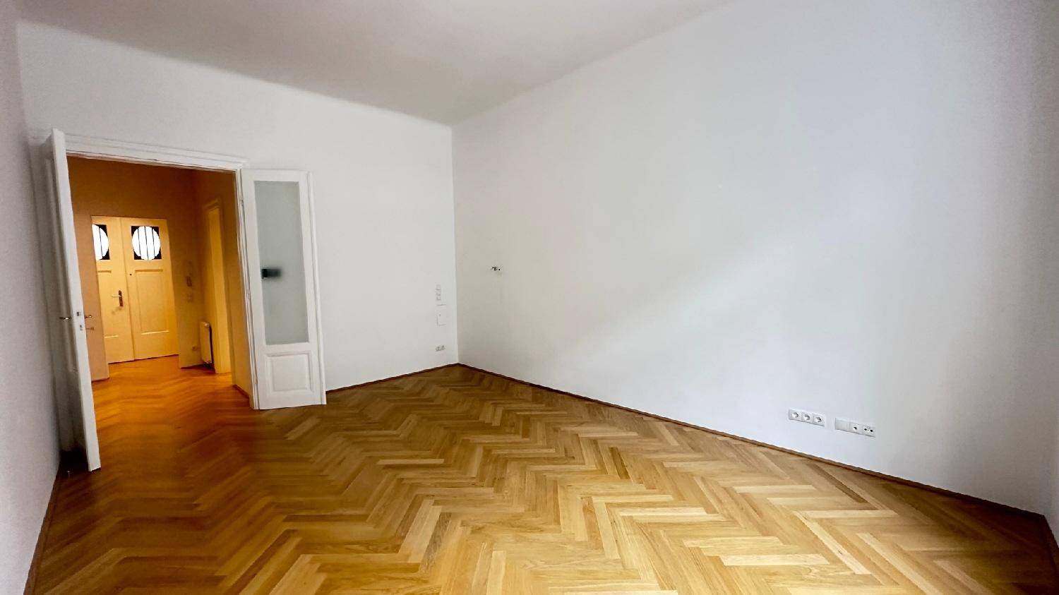 Helles, leeres Zimmer mit Fischgrätparkett und Zugang zu einem weiteren Raum, ideal für Wohnzwecke.
