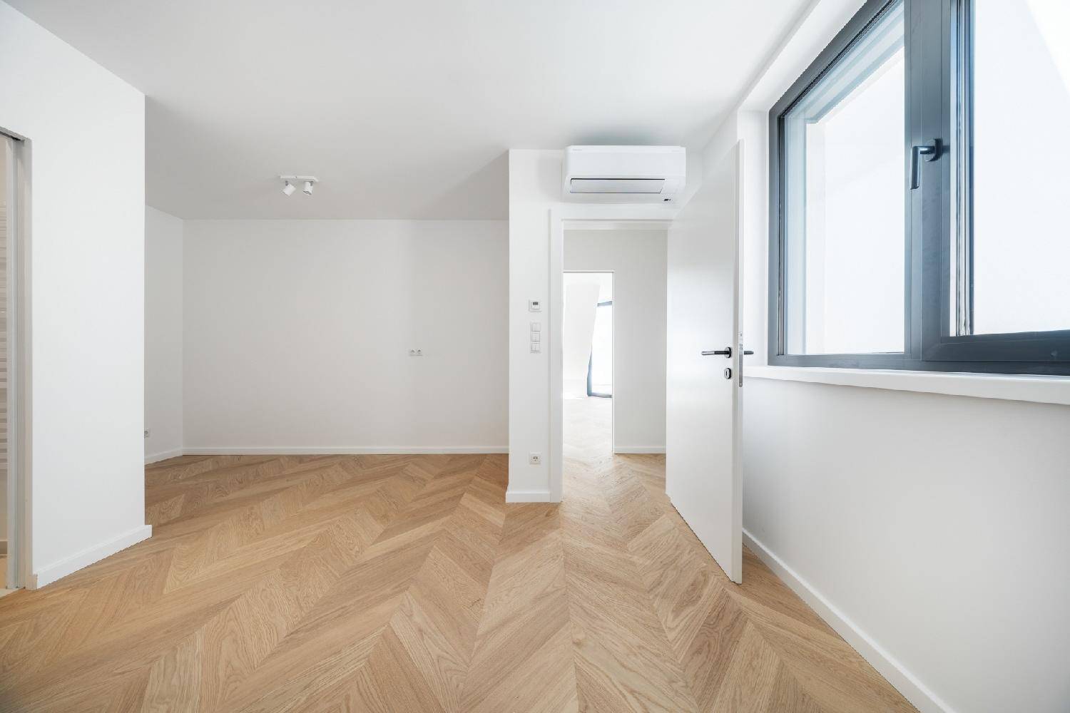Helles Schlafzimmer mit Fischgrätparkett, Klimaanlage und Fenster mit Blick nach draußen.