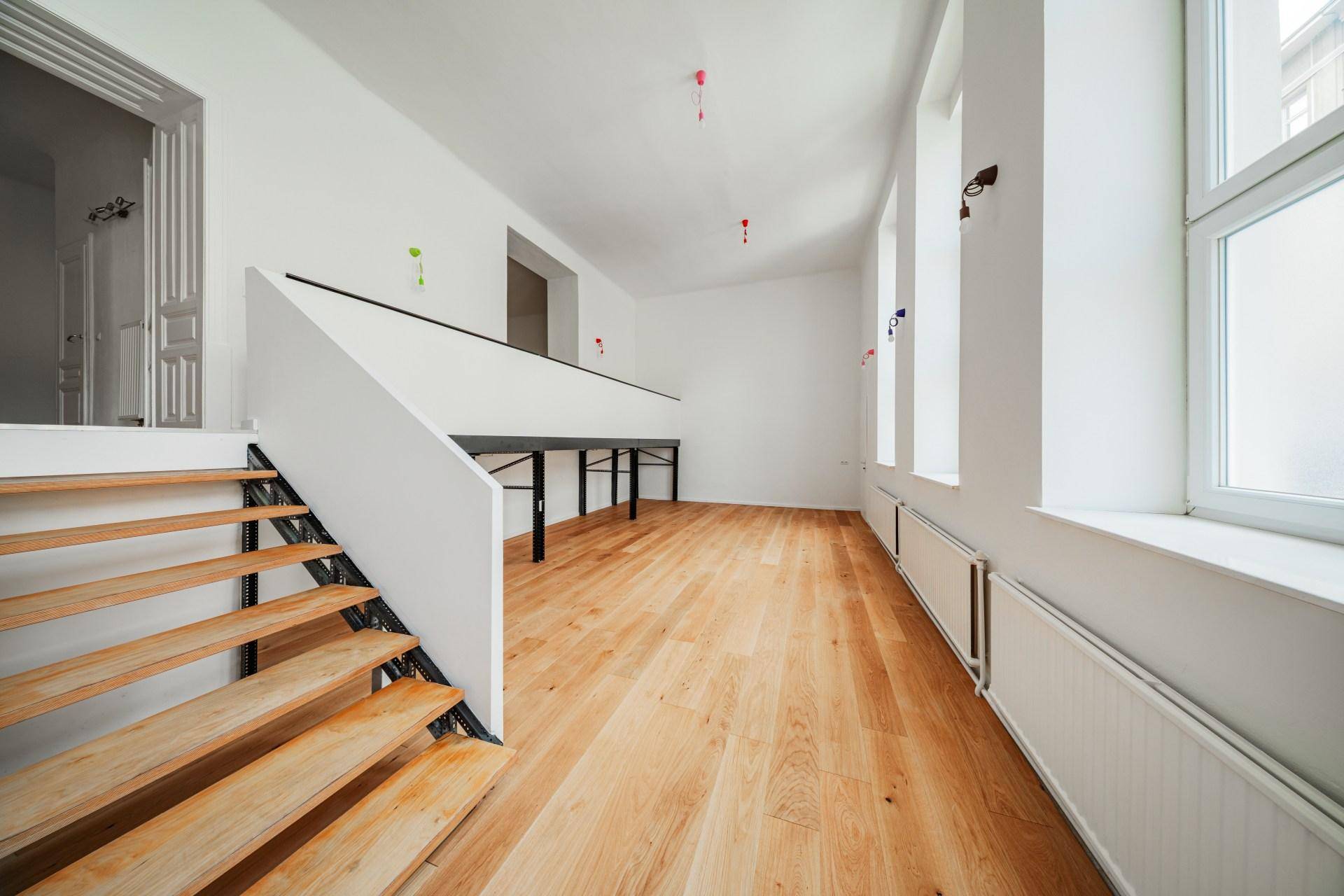 Heller Raum mit Holzboden und offener Treppe, große Fenster lassen viel Tageslicht herein.