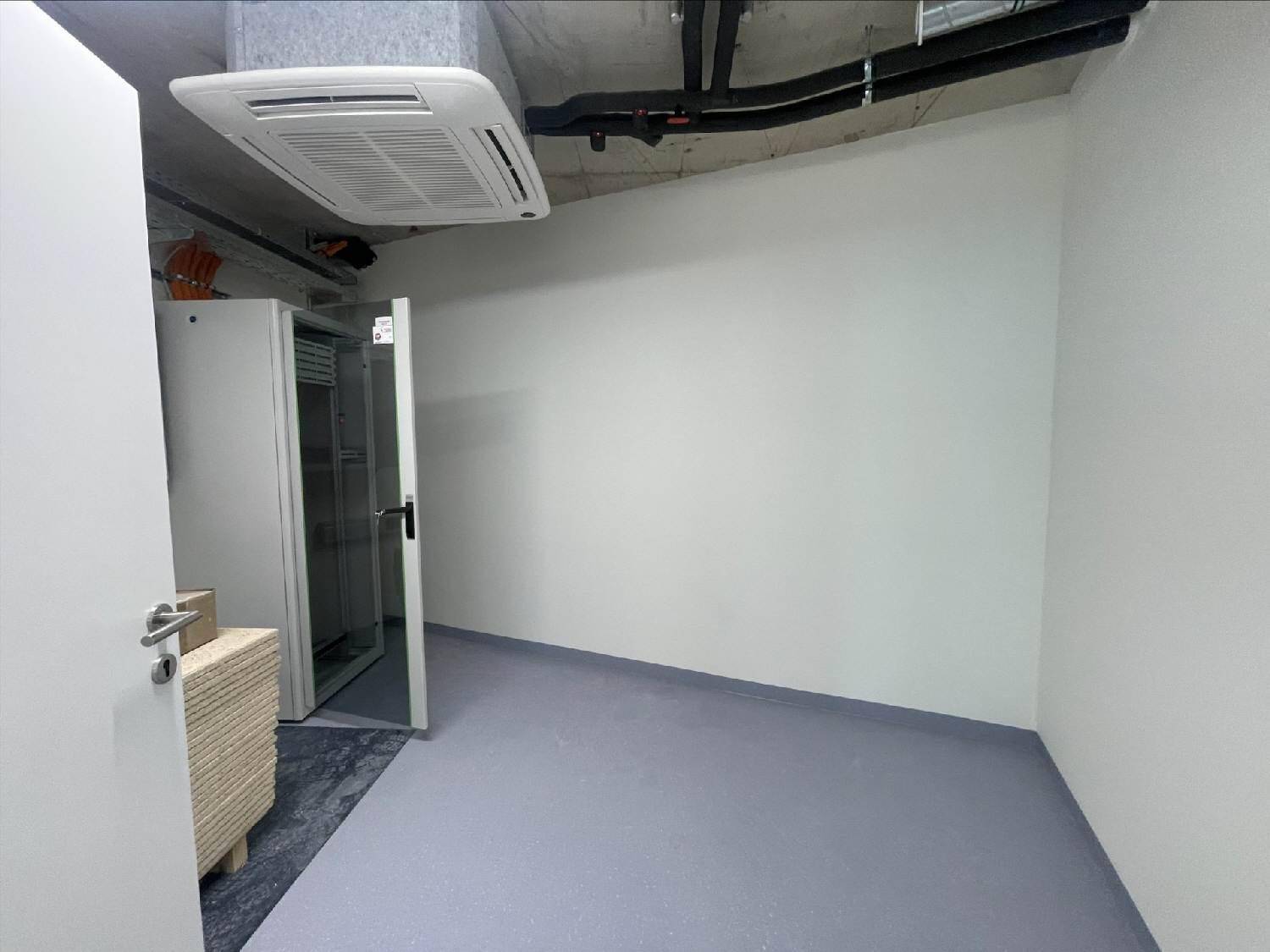 Kleiner Technikraum mit Klimaanlage an der Decke, sichtbaren Leitungen und grauem Vinylboden, funktional eingerichtet.