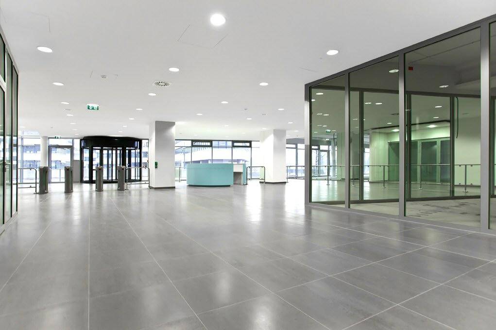 Geräumige, helle Lobby mit modernen Fliesenböden, Glaswänden und einem Empfangsbereich.