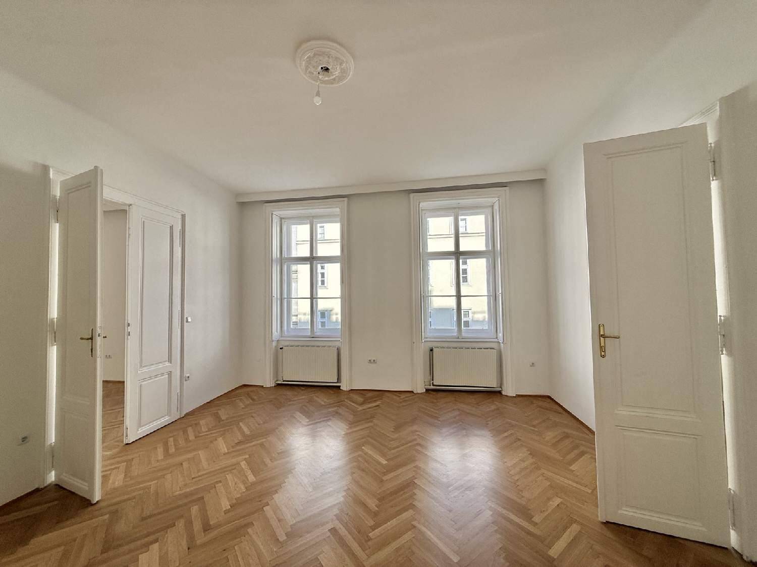 Geräumiges Zimmer mit klassischem Fischgrätparkett, zwei großen Fenstern und einer Stuckrosette an der Decke.
