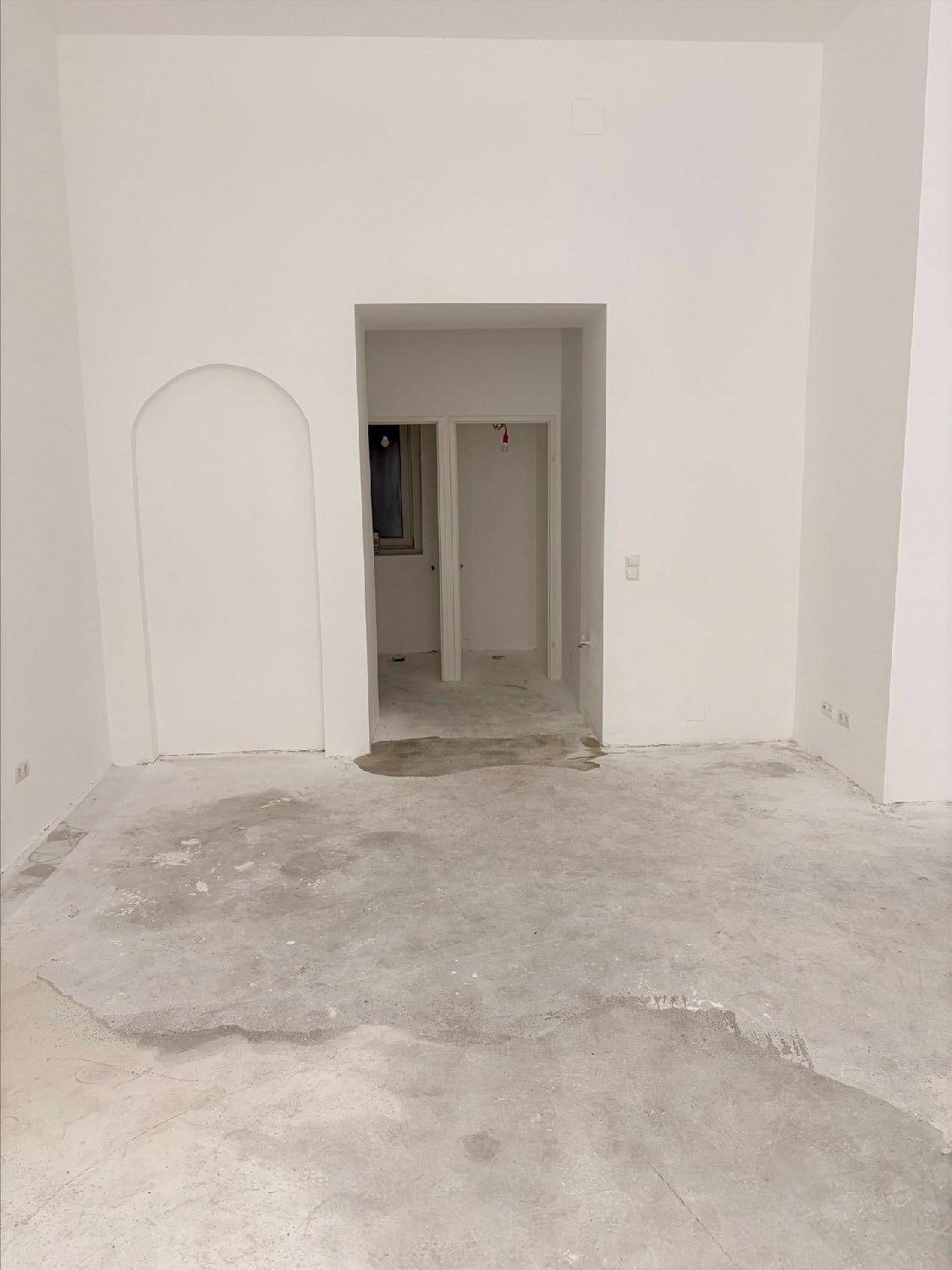 Geräumiger, leerer Raum mit weißen Wänden und Betonboden, mit einem Durchgang zu angrenzenden Bereichen.