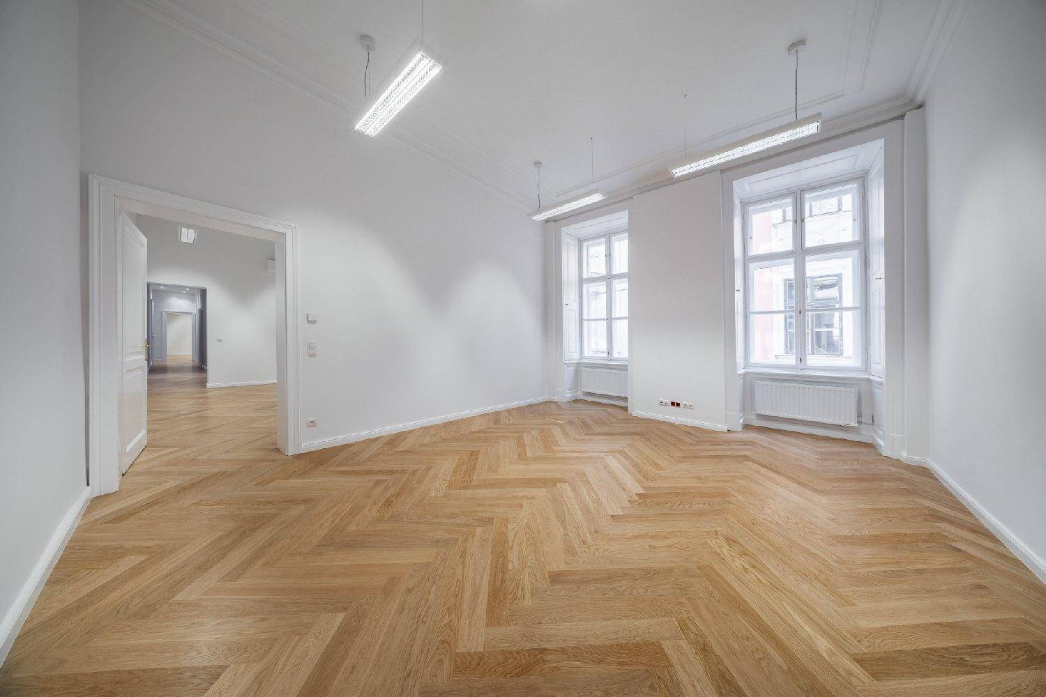 Leerer, heller Raum mit Fischgrätparkett, hohen Fenstern und modernen Deckenleuchten.