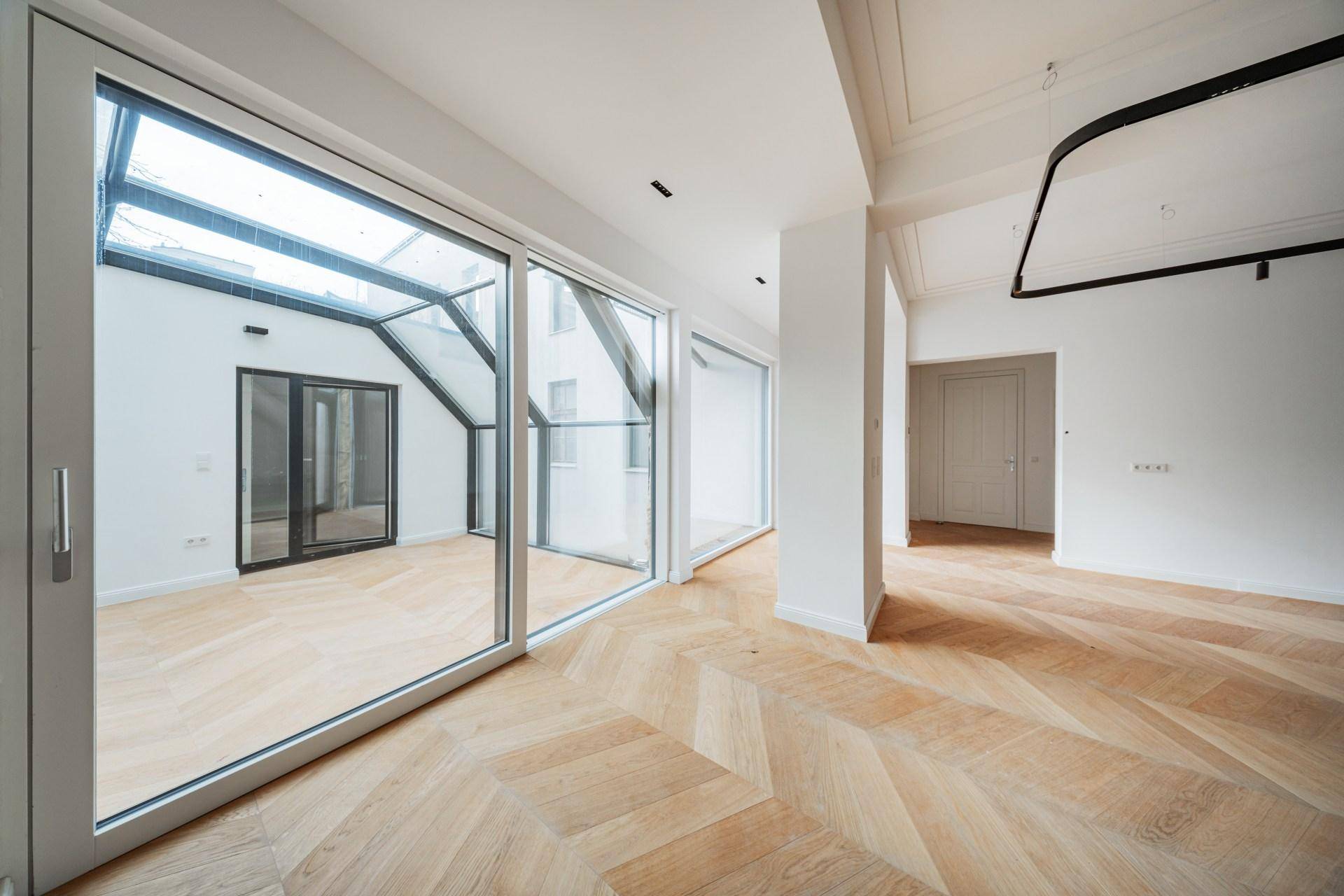Geräumiges Zimmer mit Fischgrätparkett und Glasfront, die zu einem Wintergarten oder Innenhof führt.