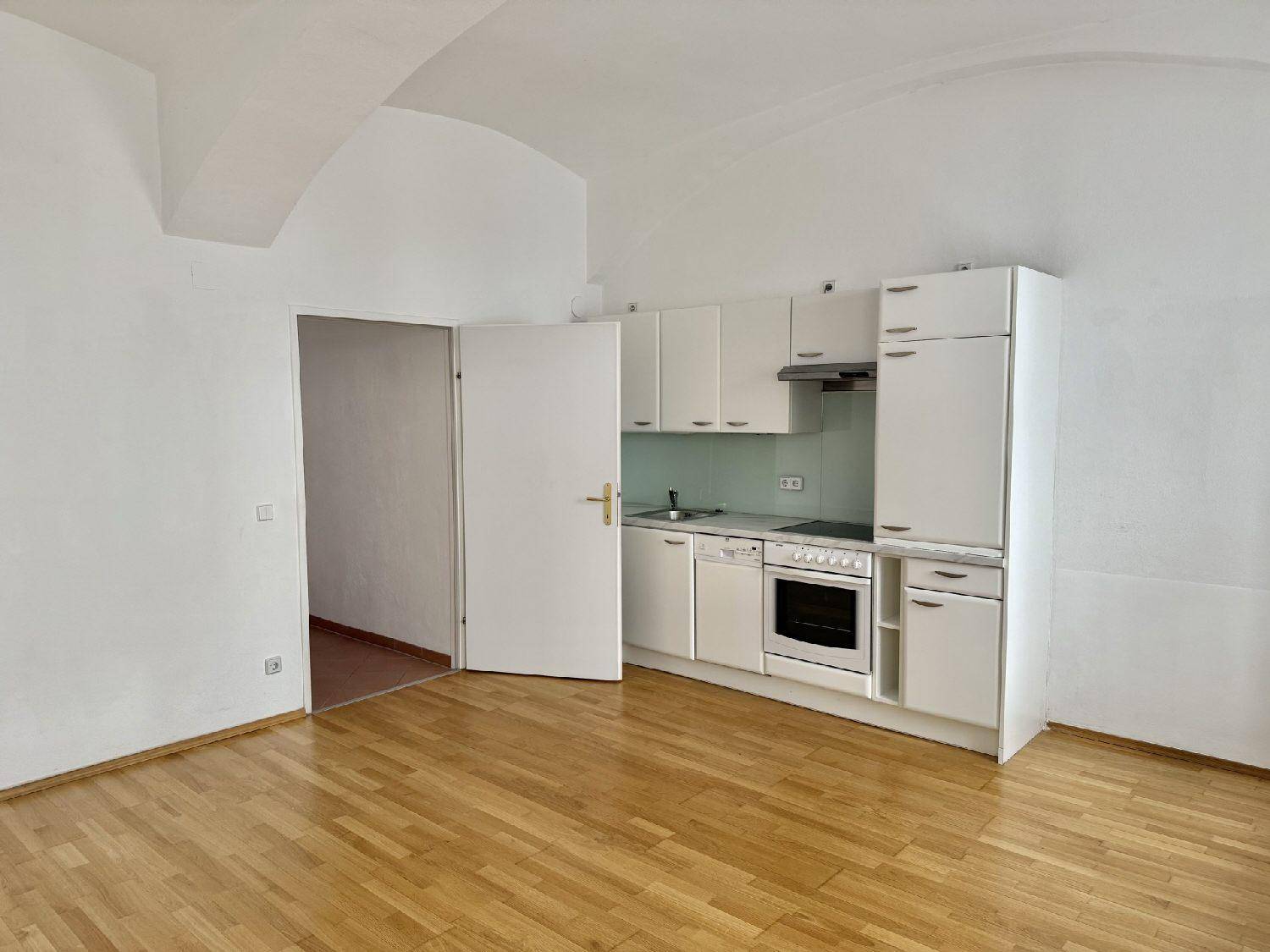 Heller Raum mit integrierter Küchenzeile, Holzboden und Zugang zu einem weiteren Zimmer.