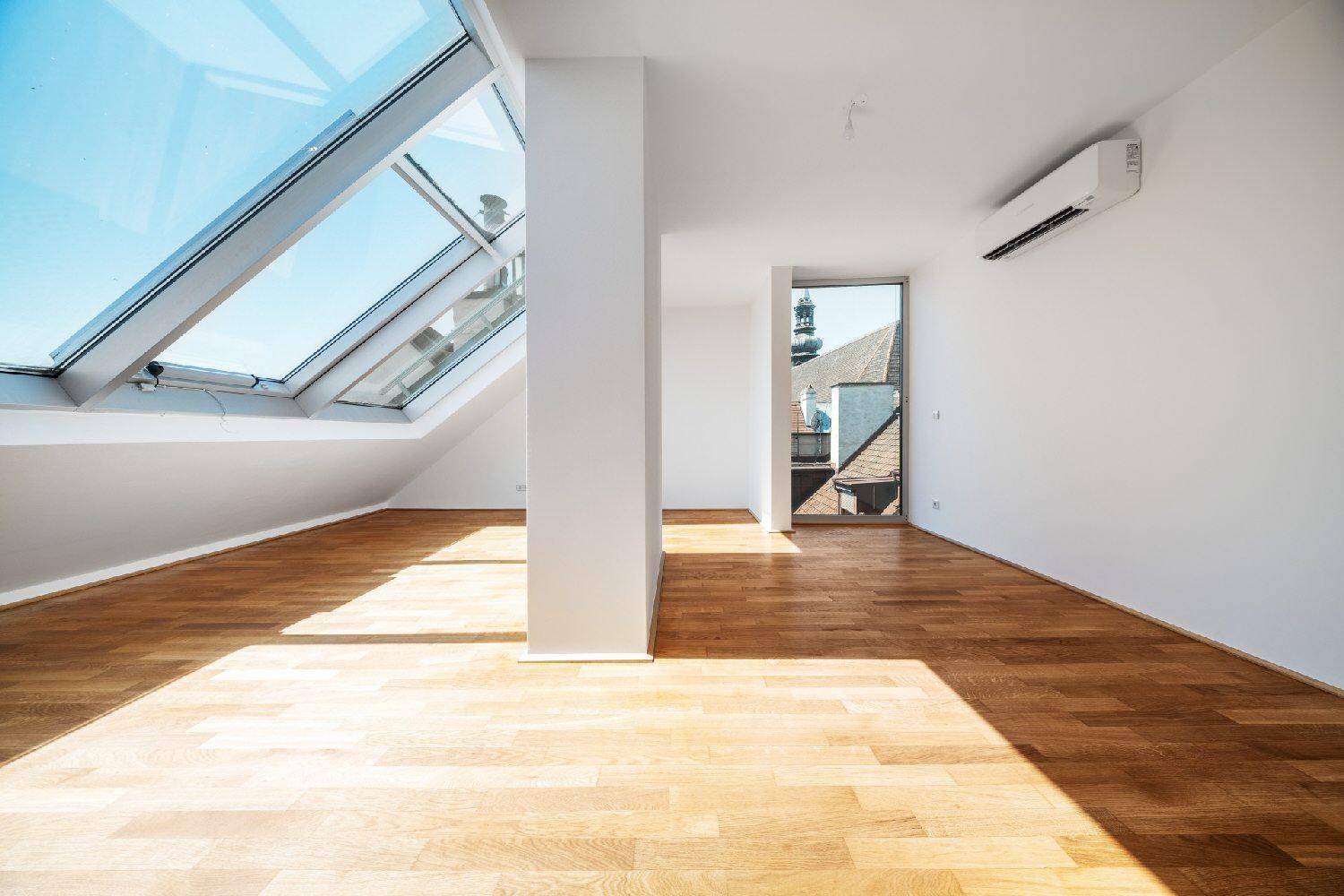 Heller Raum mit Holzboden, Dachfenstern und Zugang zu einem Außenbereich mit Kirchturmblick.