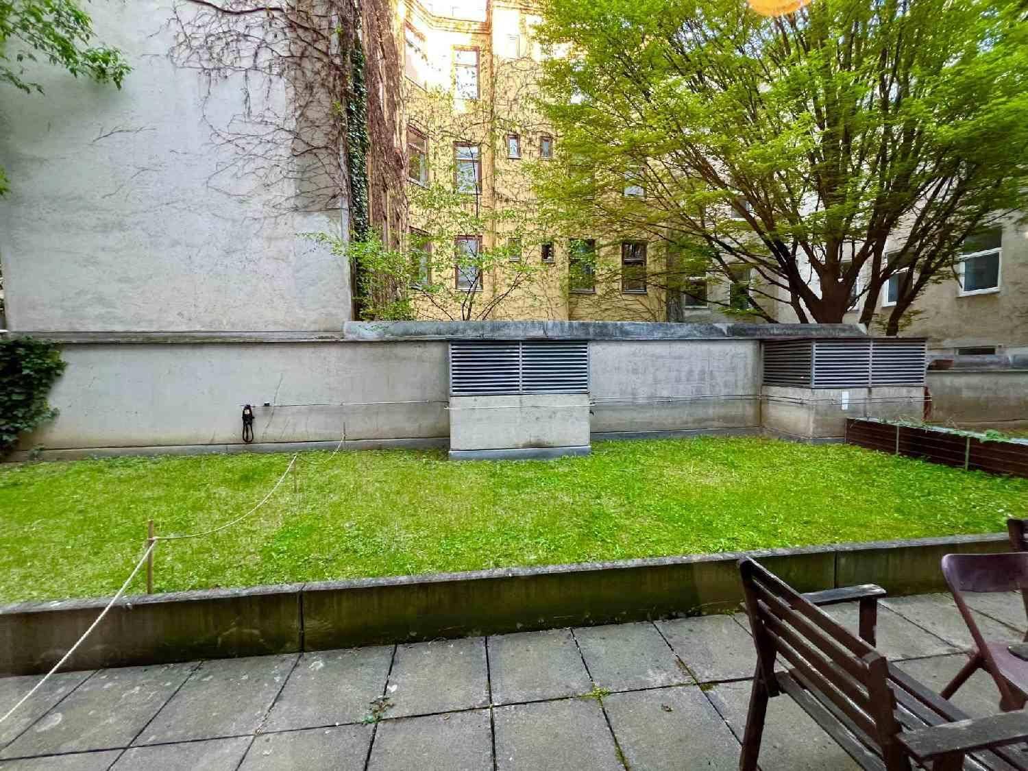 Gepflegter Gartenbereich mit Rasenfläche und gepflasterter Terrasse, umgeben von Gebäuden und Bäumen.