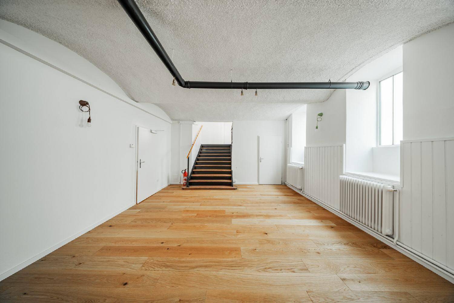 Großer, heller Raum mit Holzboden, sichtbaren Rohren und Treppe im Hintergrund, vielseitig nutzbar.