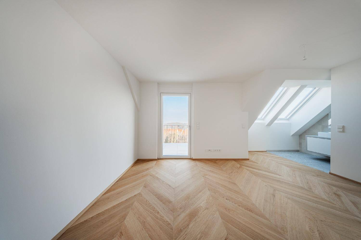 Helles Zimmer mit Dachschräge, Parkettboden und Zugang zu einem kleinen Balkon.