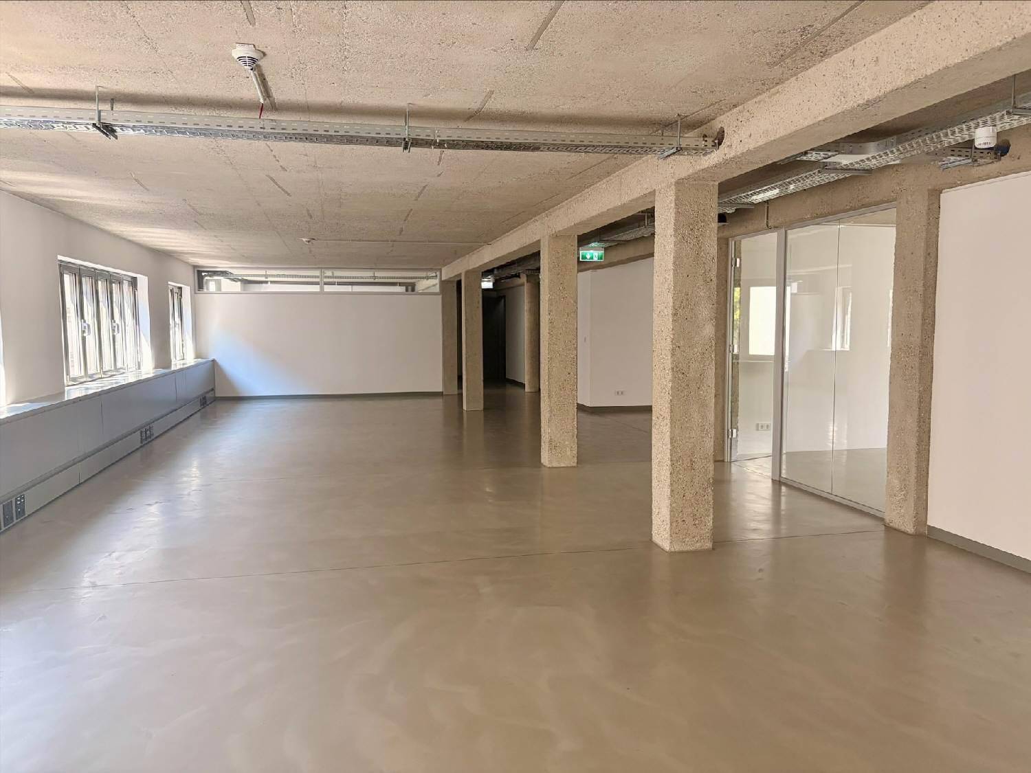 Heller Büroraum mit Betonboden, Glaswänden und sichtbaren Installationen an der Decke.