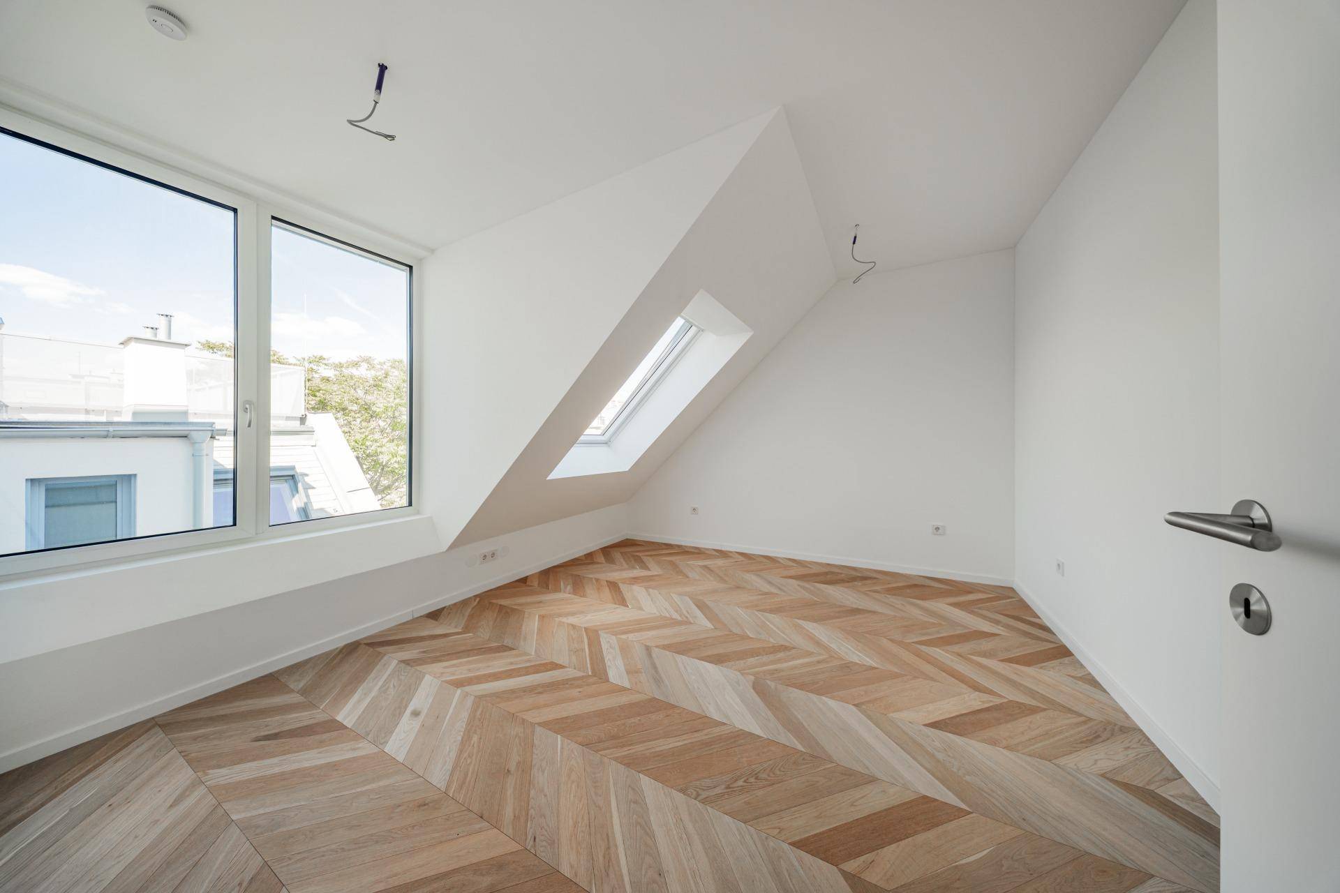 Helles Schlafzimmer mit Fischgrätparkett, großem Fenster und Dachfenster in der Schräge.