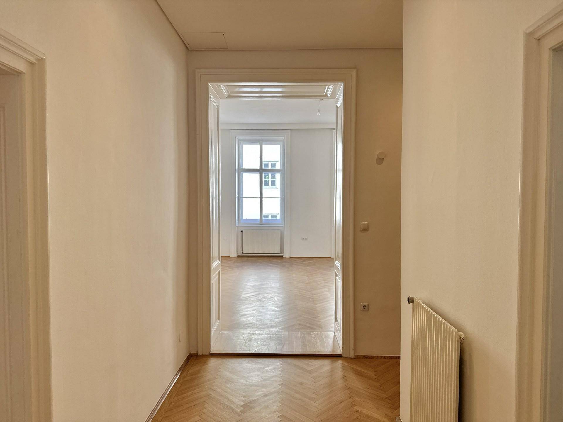 Blick vom Flur in ein weiteres helles Zimmer mit Fischgrätparkett und einem großen Fenster.
