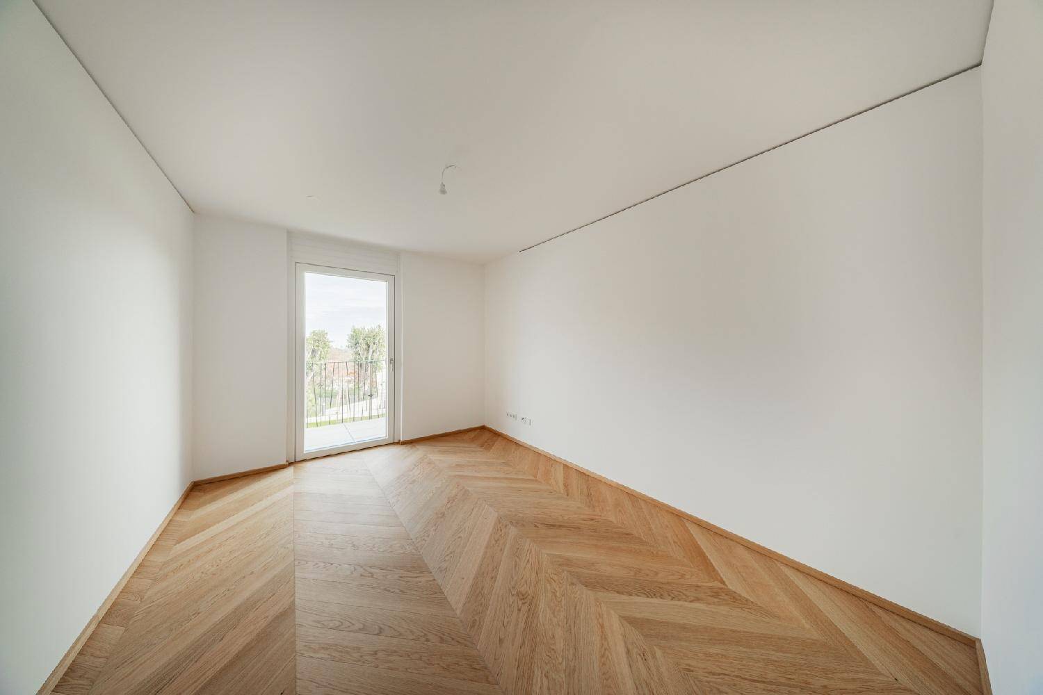 Leeres Zimmer mit Fischgrätparkett und einem Fenster mit Blick ins Grüne.