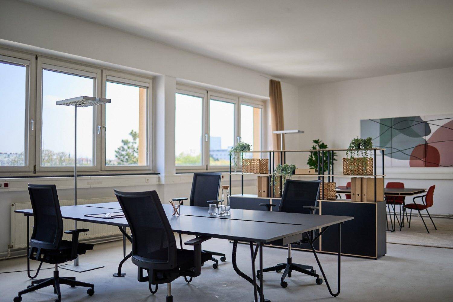 Helles Großraumbüro mit mehreren Arbeitsplätzen, großen Fenstern und moderner Einrichtung.