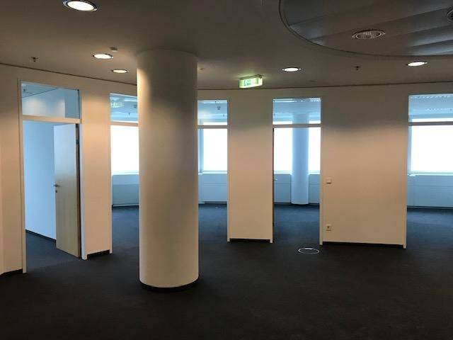Offener Bürobereich mit dunklem Teppichboden, Säulen und Zugang zu weiteren Räumen.