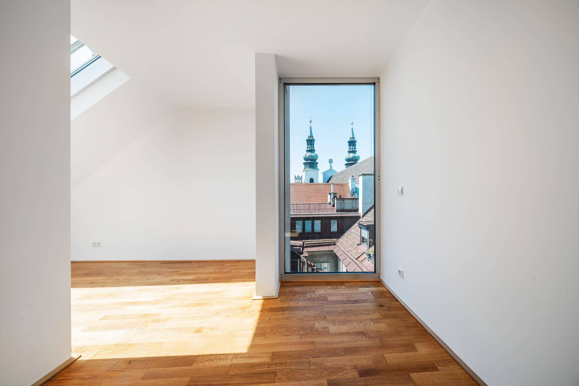 Heller Raum mit Holzboden und hohem Fenster, das einen weiten Blick über die Dächer der Stadt bietet.