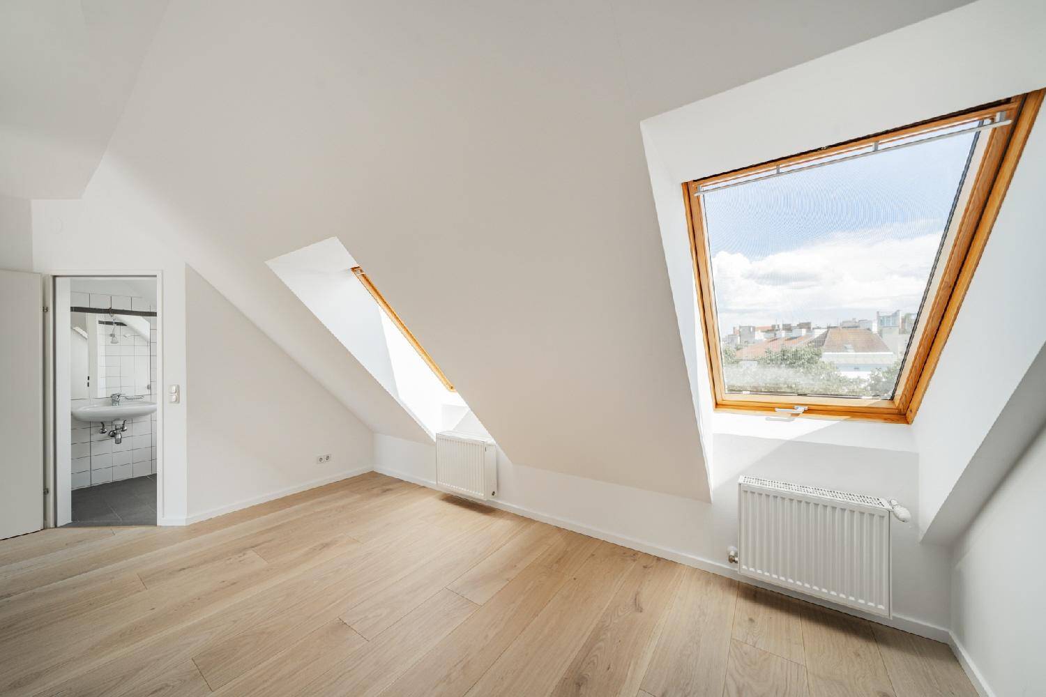 Helles Dachzimmer mit schrägen Wänden, Holzboden und Dachfenstern, die einen Blick über die Dächer bieten.
