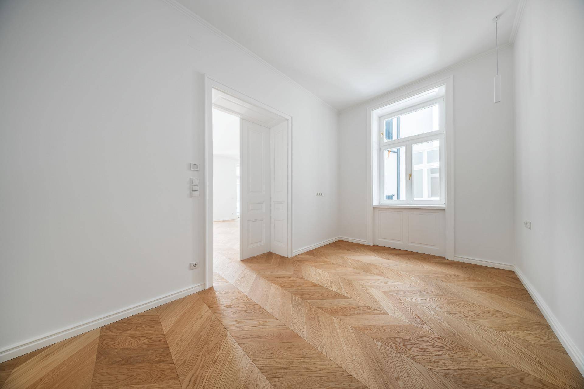 Leeres Zimmer mit hellem Fischgrätparkett, weißen Wänden und einem großen Fenster.