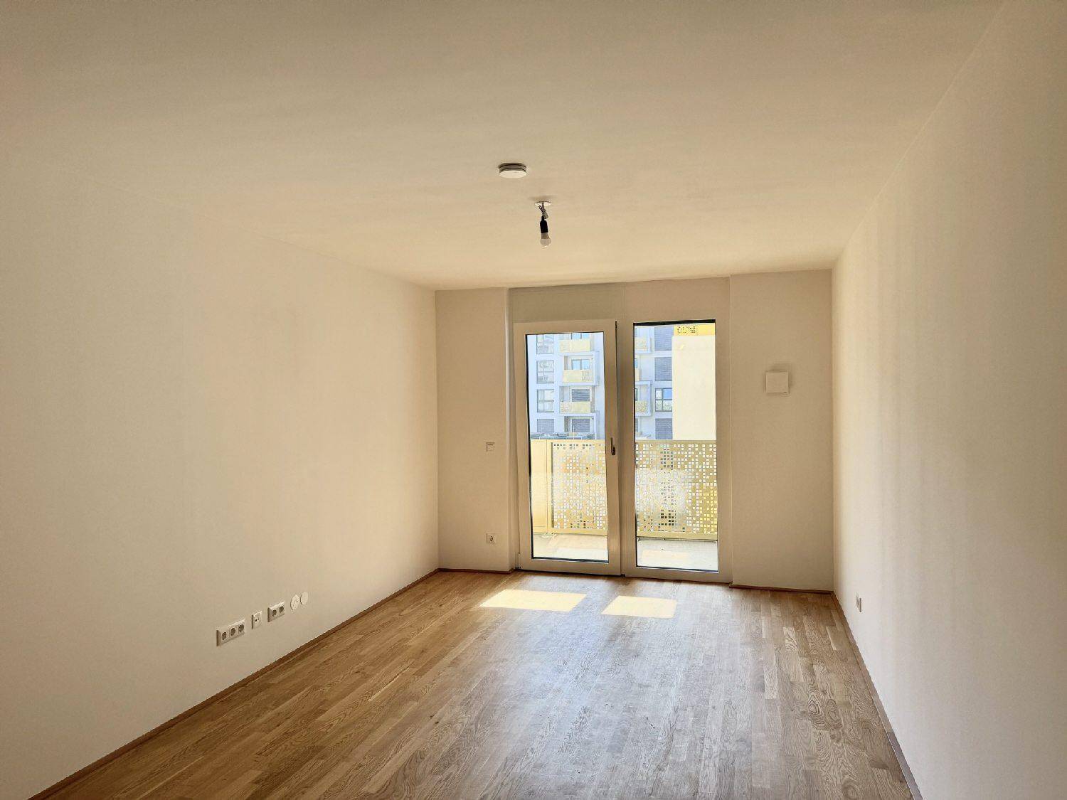 Helles, leeres Zimmer mit Parkettboden und Zugang zu einem Balkon.