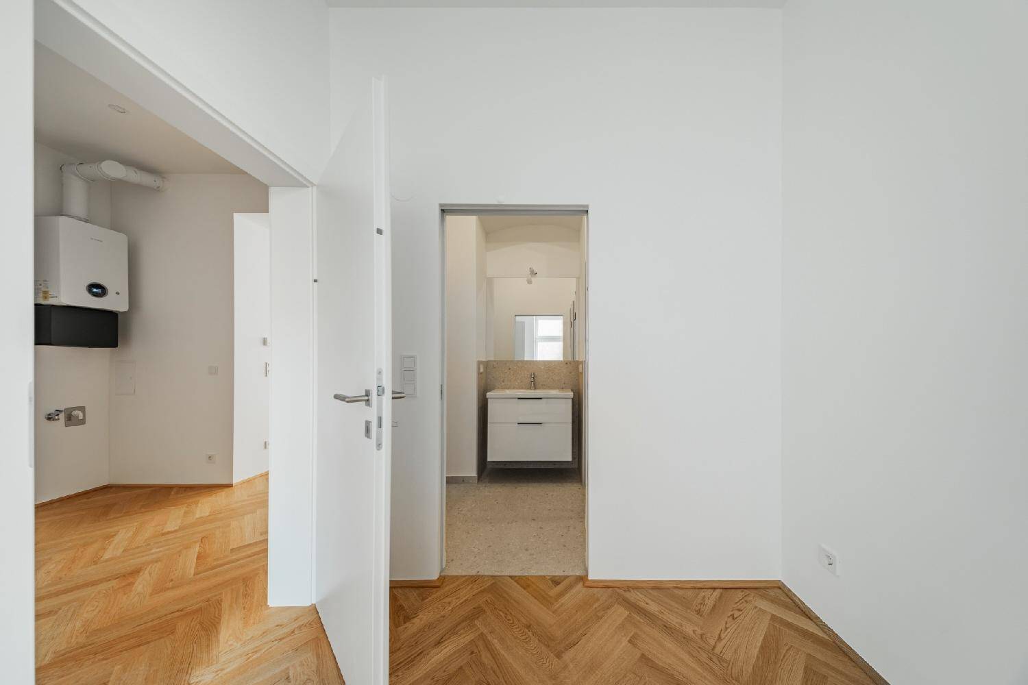 Flur mit Fischgrätparkett und Blick in ein modernes Badezimmer mit Waschtisch.