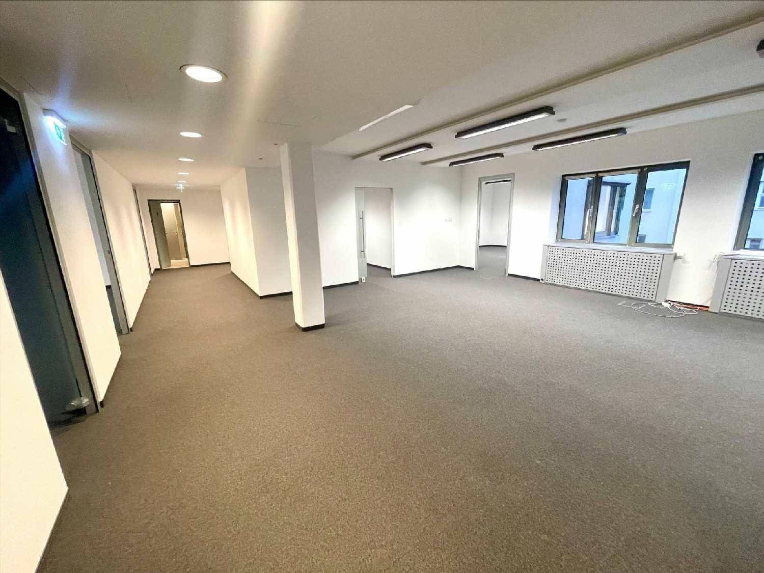 Langer Bürobereich mit Teppichboden, Säulen und Fenstern, die Tageslicht hereinlassen.