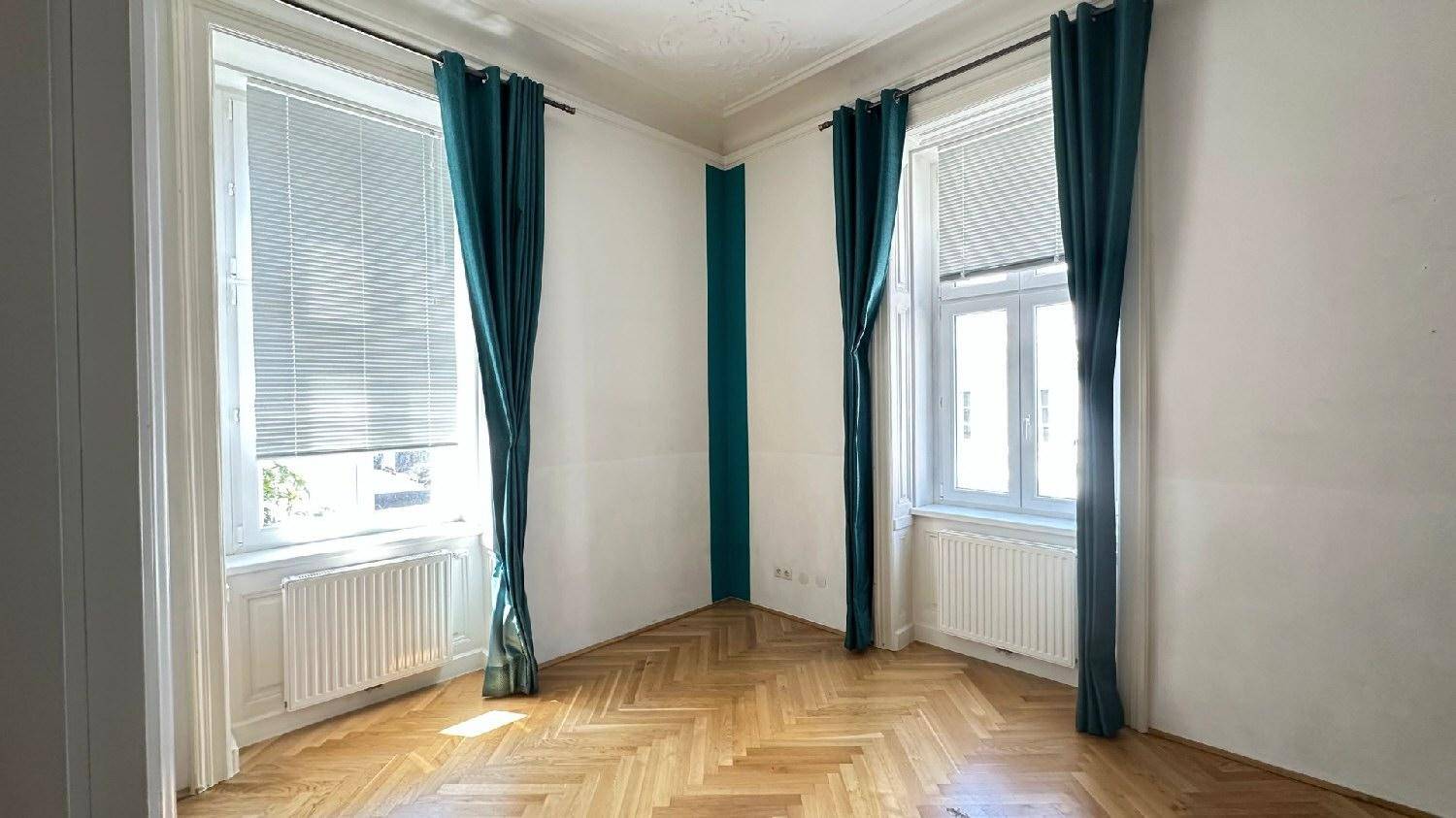 Leeres Zimmer mit Fischgrätparkett, hohen Fenstern und blauen Vorhängen.
