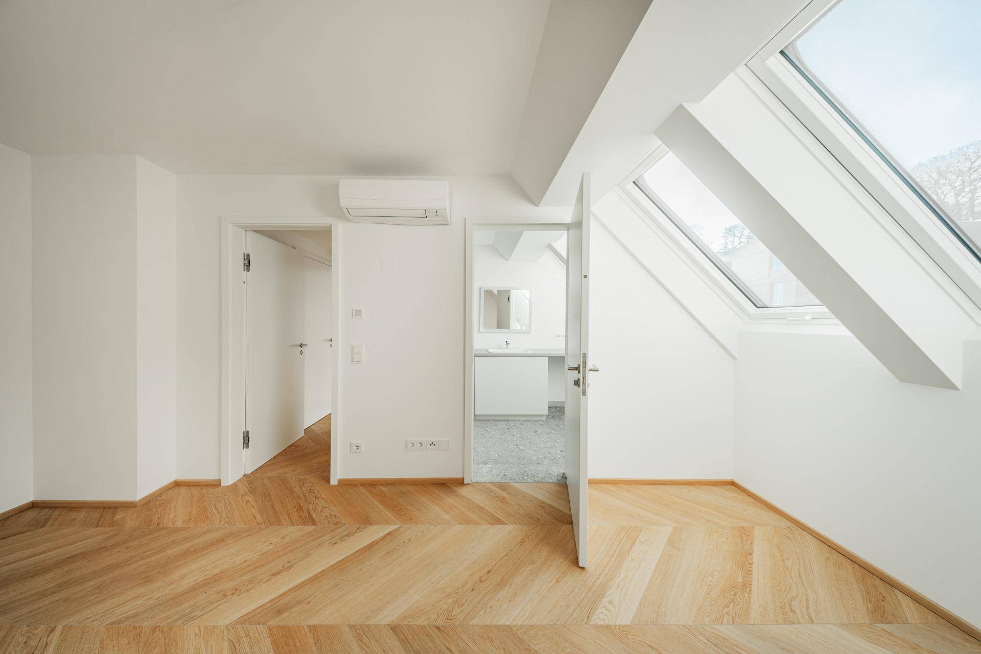 Heller Raum mit Fischgrätparkett und Zugang zu einem modernen Badezimmer mit Dachfenster.