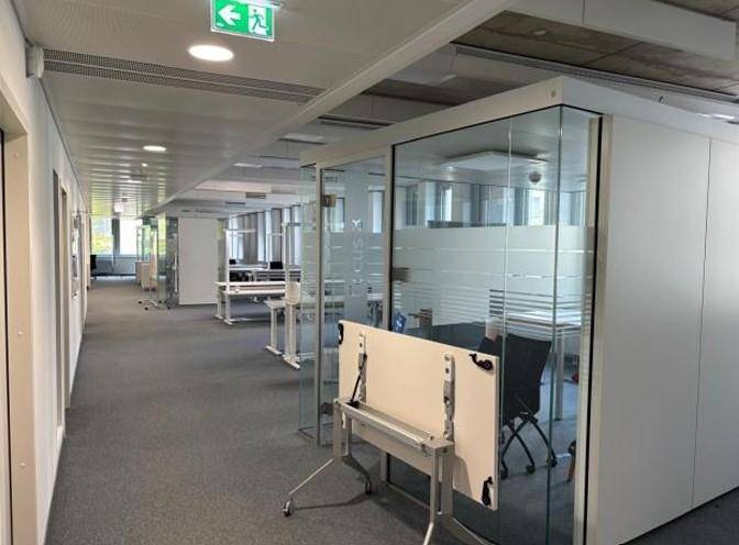 Heller Büroflur mit Teppichboden und modernen Glaswänden, die zu den einzelnen Büros führen.