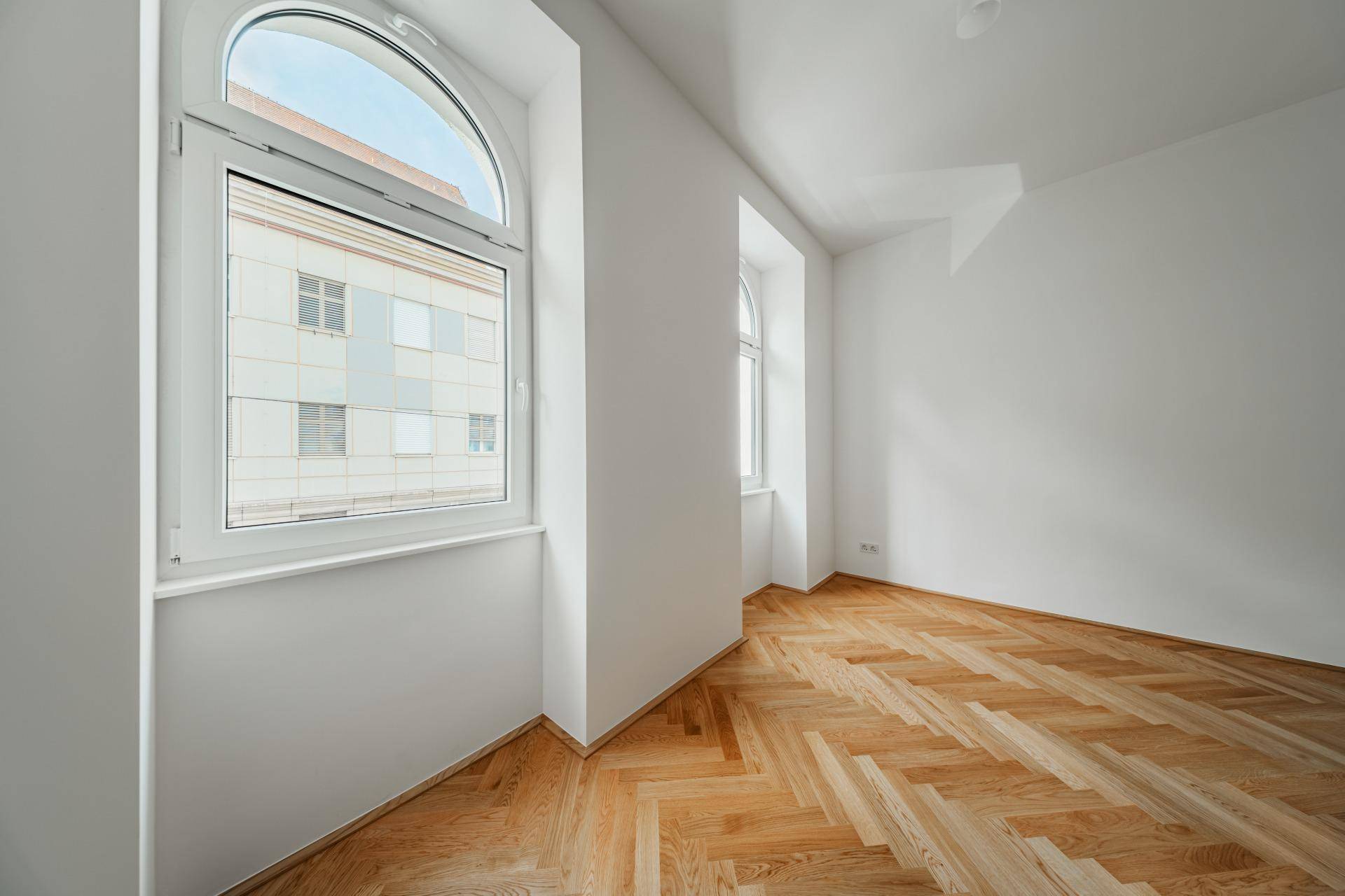 Heller Raum mit Fischgrätparkett und großen Bogenfenstern, die viel Tageslicht hereinlassen.