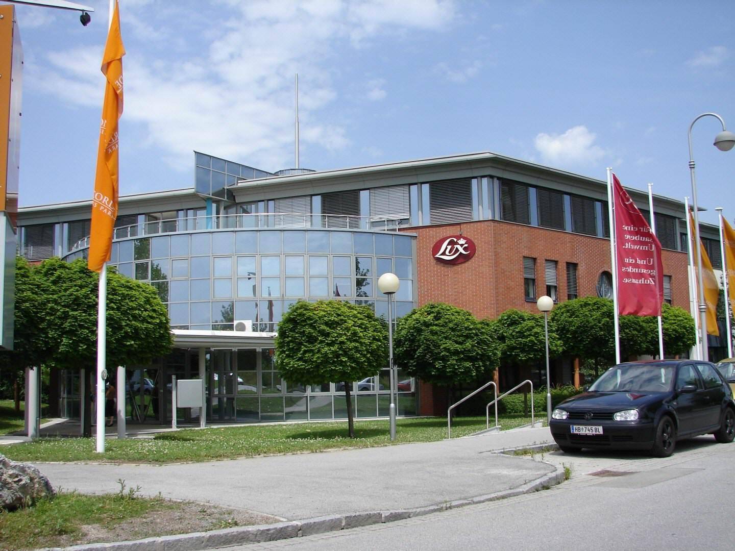 Modernes Geschäftsgebäude mit geschwungener Glasfassade und Ziegelbereichen, umgeben von Fahnen und Bäumen.