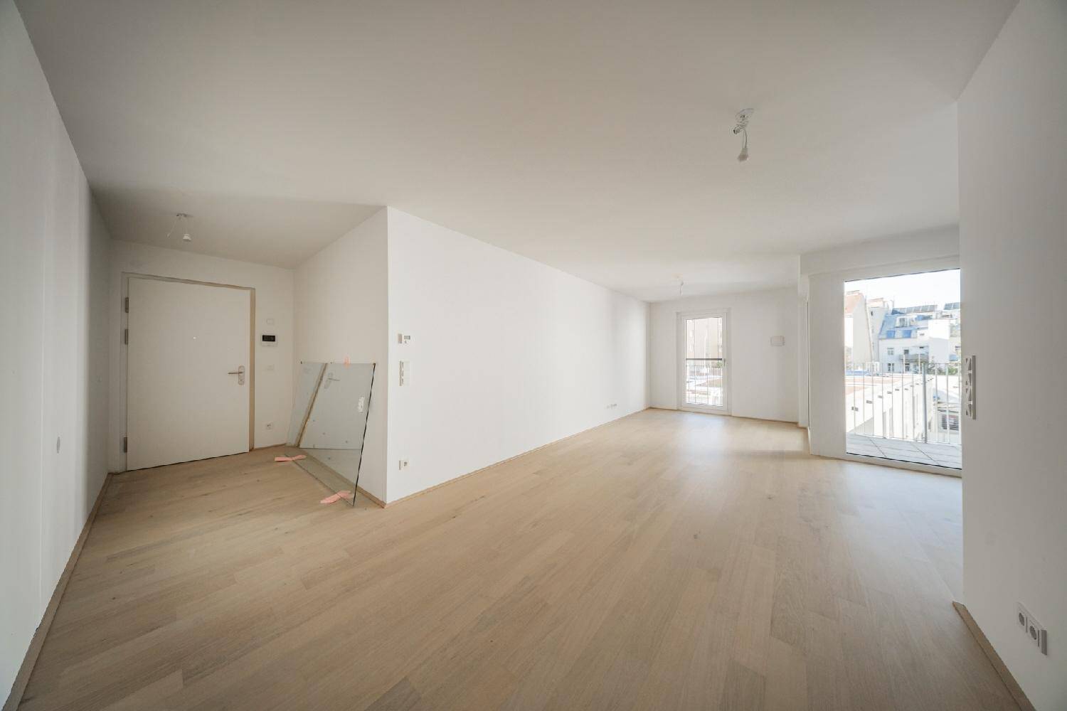Heller Wohnbereich mit Parkettboden, weiß gestrichenen Wänden und Zugang zum Balkon.
